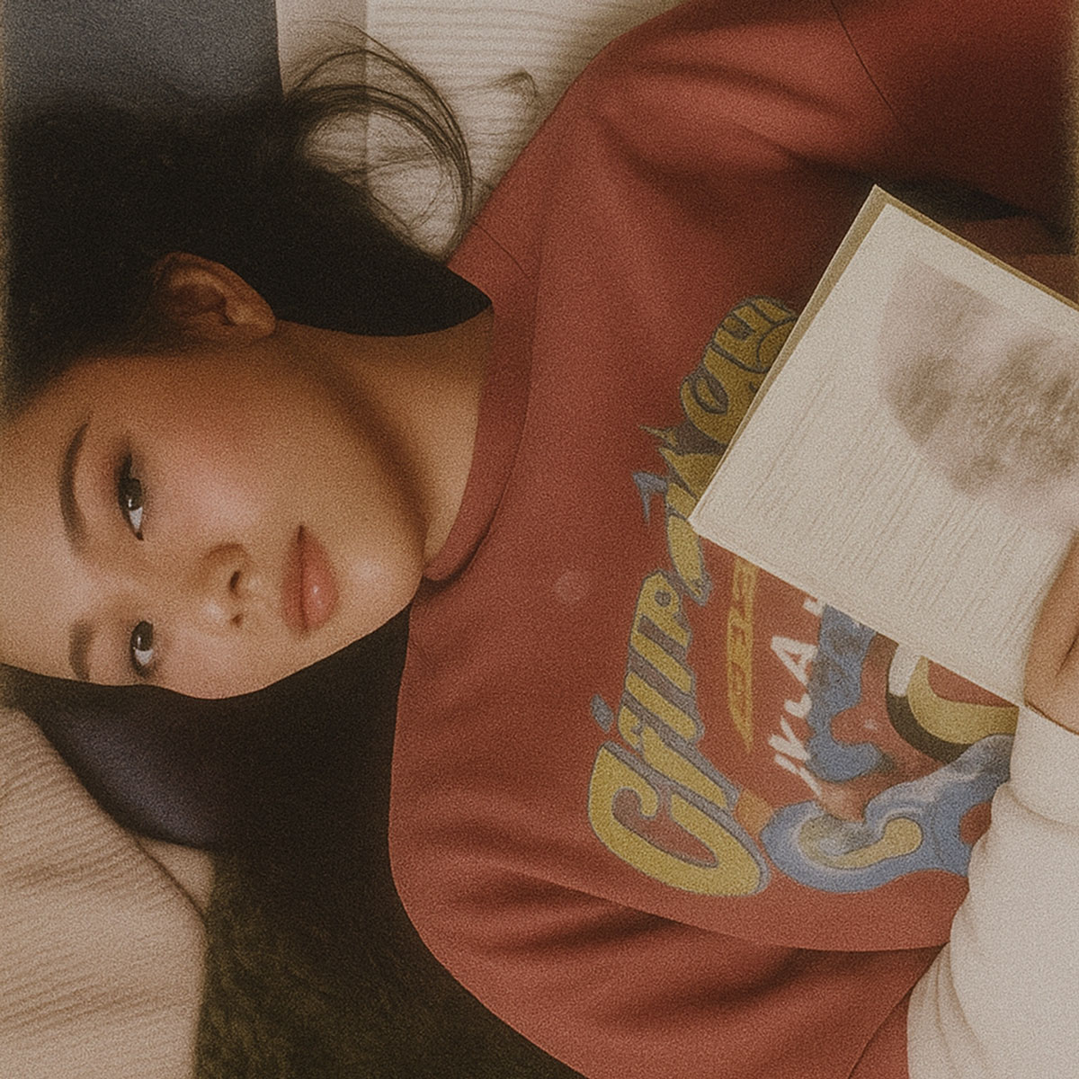 ภาพผู้หญิงสวมเสื้อสเวตเชิ้ตสีแดงลายน่ารัก นอนเอนอ่านหนังสือบนพื้นห้องในบรรยากาศอบอุ่น แสงนวลและโทนสีวินเทจ ถ่ายทอดความรู้สึกสบายและมีสไตล์แบบแบรนด์ kangpla